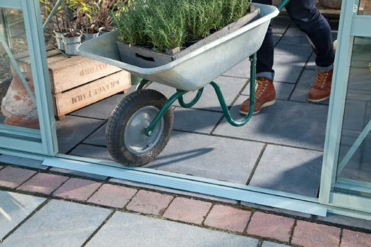 A person in boots pushes a wheelbarrow with plants through an open Rhino Greenhouse door on a paved patio. Two wooden crates are visible on the left.