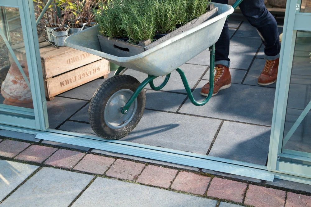 A wheelbarrow carries potted plants, pushed by someone wearing boots, inside a Rhino Greenhouse. Nearby are wooden crates on a tiled floor.