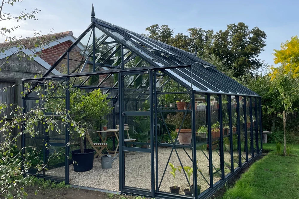 A large glass and metal Rhino Greenhouse houses various potted plants and a small wooden table set, situated in a grassy garden area, with trees and a brick building nearby.