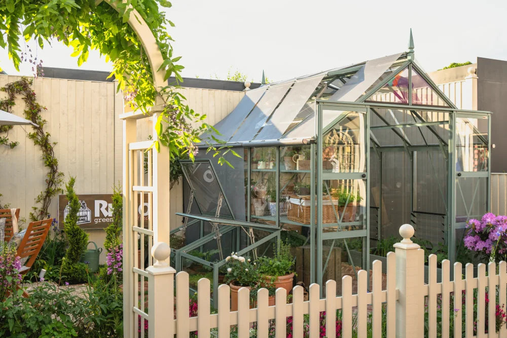 A Rhino Greenhouse with open windows houses potted plants and gardening tools, surrounded by a white fence and lush garden. A sign reads Rhino Rhino Greenhouses on a wooden wall.