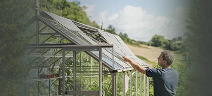 A person adjusts a vent on a glass-paneled Rhino Greenhouse in a lush, green garden setting with rolling hills and a partly cloudy sky in the background.