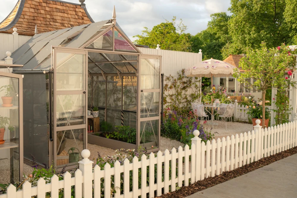 A Rhino Greenhouse with open doors showcases plants inside, surrounded by a white picket fence. Adjacent, a garden area features a patio table, umbrella, and flowering plants.