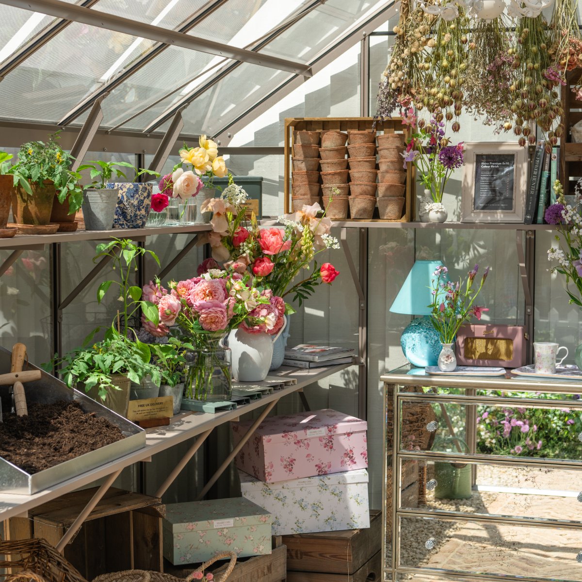 Flowers in vases are displayed on Rhino Greenhouse shelves, surrounded by potted plants, dried herbs, and gardening tools. Gift boxes and decorative items enhance the sunlit, colorful setting.