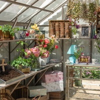 Flowers in vases are displayed on Rhino Greenhouse shelves, surrounded by potted plants, dried herbs, and gardening tools. Gift boxes and decorative items enhance the sunlit, colorful setting.