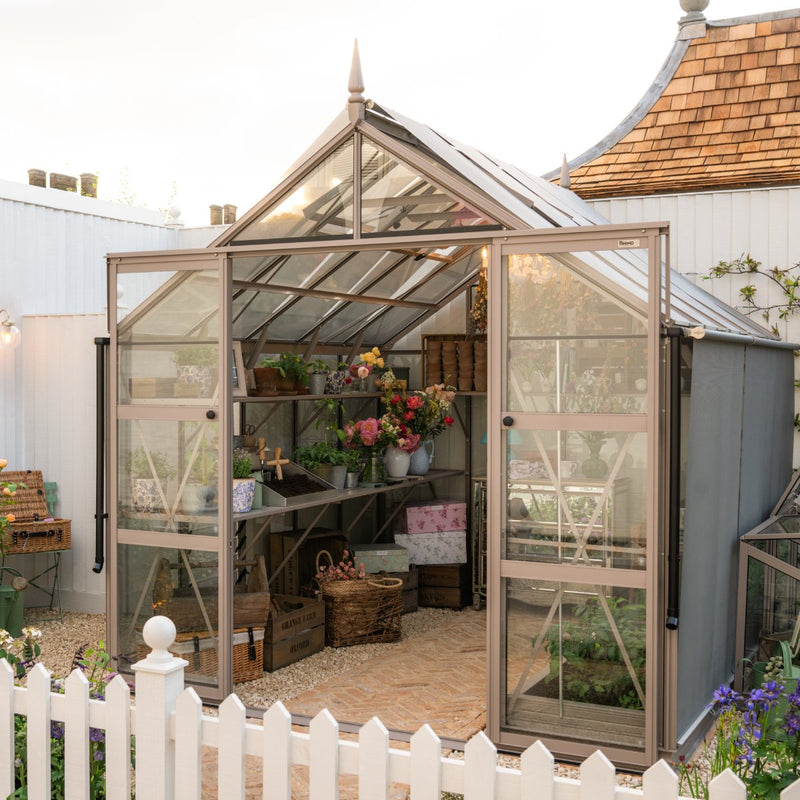 A glass Rhino Greenhouse houses potted plants and flowers on metal shelves. It's surrounded by a brick path and white picket fence, with a wooden building roof partially visible behind it.