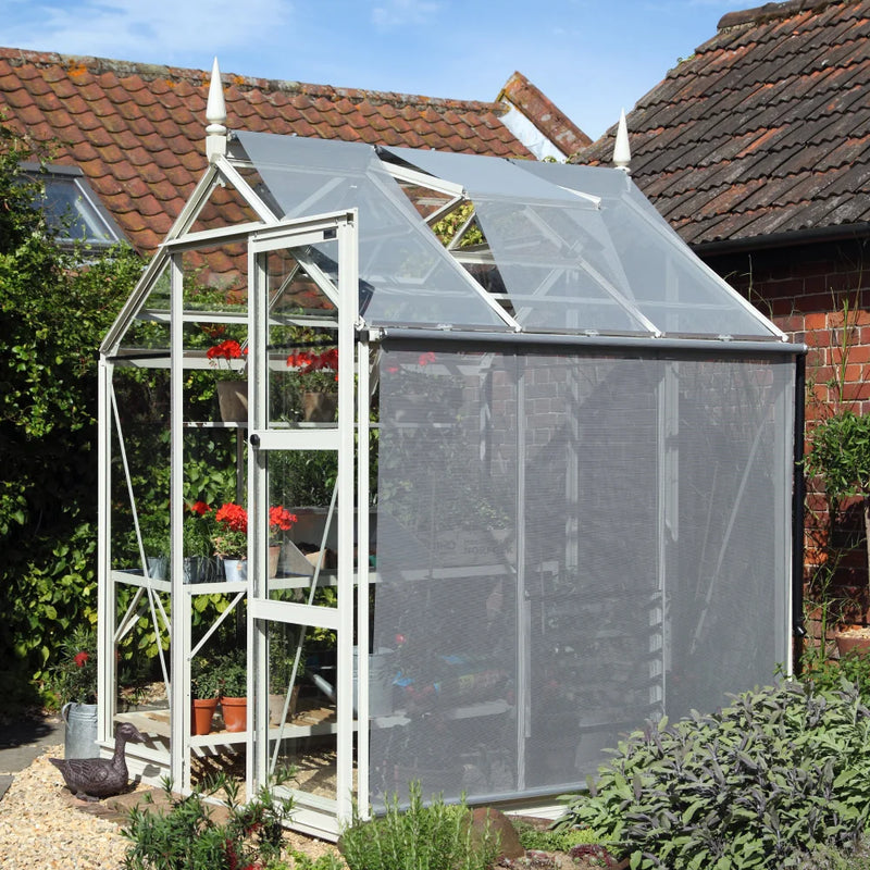 A glass Rhino Greenhouse with an open door displays vibrant red flowers in pots. It's situated in a garden with brick buildings and greenery surrounding it.