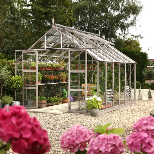A Rhino Greenhouse stands in a gravel garden, housing potted plants on shelves. Surrounding it are lush trees and vibrant pink flowers, creating a serene outdoor setting.