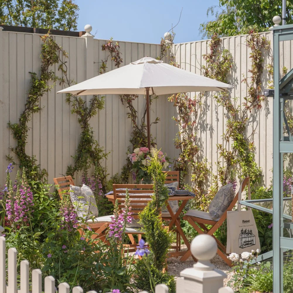 A wooden table with chairs and umbrella sits in a flower-filled garden, enclosed by trellised fencing. A tote bag labeled Rhino Rhino Greenhouses rests nearby.