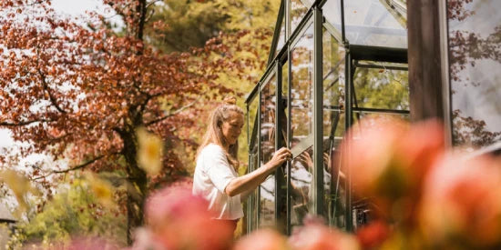 A woman is opening a Rhino Greenhouse door surrounded by blossoming flowers and lush trees, creating a serene garden setting.