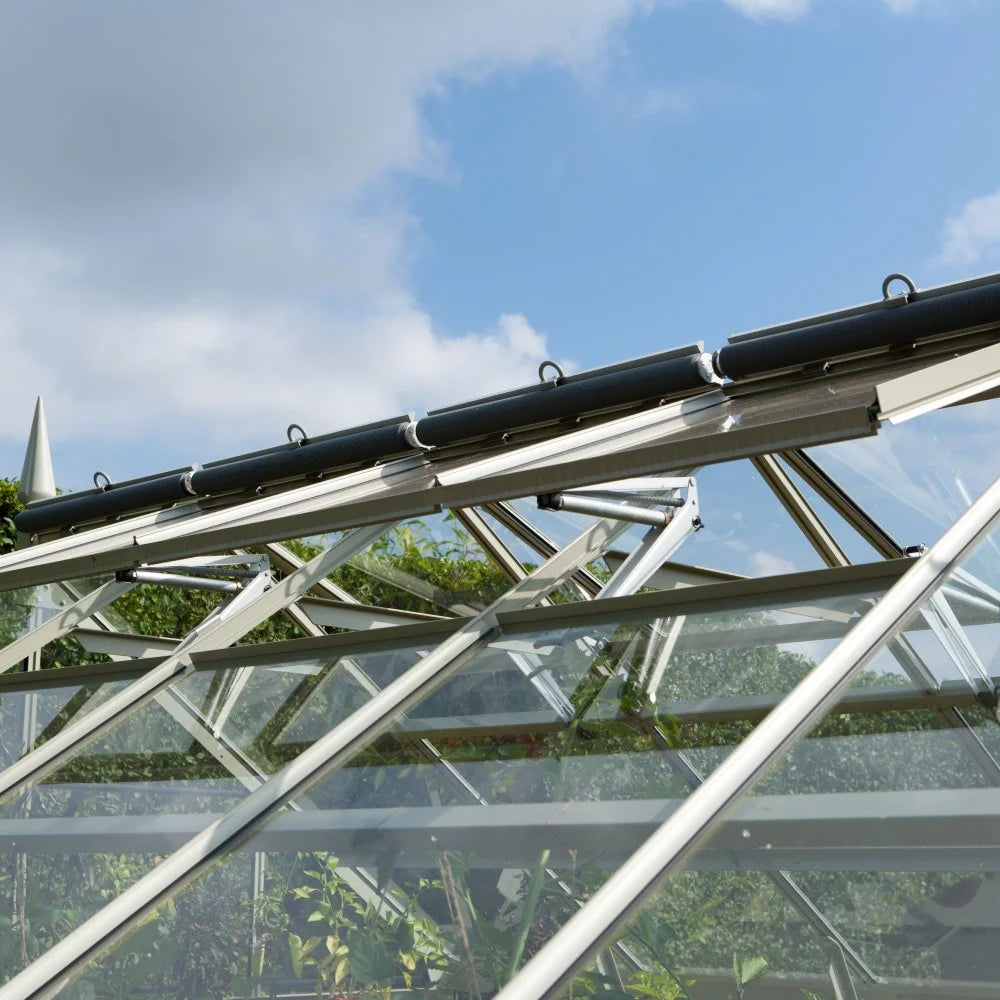 Glass Rhino Greenhouse panels form a structure with metal frames supporting them. The panels reflect sunlight against a backdrop of a blue sky with scattered clouds.